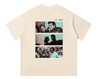 Könnte beinhalten: Ein cremefarbenes Kurzarm-T-Shirt mit einem Grafikdruck auf der Rückseite. Der Druck besteht aus drei übereinander angeordneten rechteckigen Feldern mit Szenen aus dem Film Grease, wobei der Text CASA ROMANO über dem obersten Feld steht.