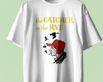 Könnte beinhalten: Ein weißes Kurzarm-T-Shirt mit dem gelben Schriftzug "the CATCHER in the RYE" über einer grafischen Illustration einer Person mit roter Jagdmütze, die eine Zigarette raucht, wobei mehrere schwarze Vogelsilhouetten über dem Kopf zu sehen sind.