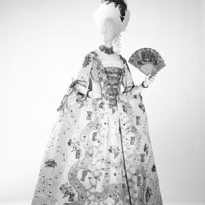 May include: A white and silver dress with a floral pattern. The dress has a fitted bodice and a full skirt. The dress is being worn by a mannequin with a white wig and a fan.