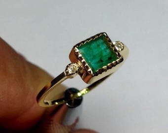 May include: A gold ring with a square emerald gemstone and two small diamonds. The emerald is set in a gold bezel with a decorative edge.