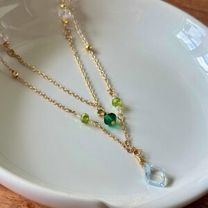 Può includere: Una delicata collana dorata a tre fili. La collana presenta perline di cristallo verdi e trasparenti, con un pendente a forma di diamante azzurro in basso. La collana è esposta su una superficie bianca.