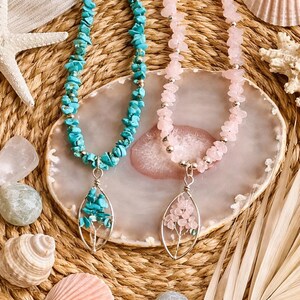 Puede incluir: Dos collares de piedras preciosas con colgantes. Uno presenta chips de turquesa y un colgante de turquesa. El otro tiene chips de piedras rosas y un colgante de flores rosas. Los collares se exhiben con conchas marinas y piedras.
