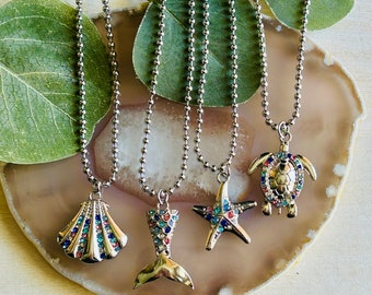May include: Five silver necklaces with different sea-themed charms. The charms are a seashell, a mermaid tail, a starfish, and a turtle. The charms are all decorated with colorful rhinestones.
