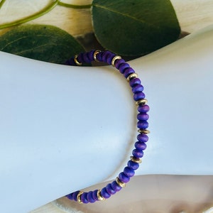 Puede incluir: Un brazalete de cuentas moradas y doradas. El brazalete está hecho de pequeñas cuentas redondas que están enhebradas en un cordón delgado. Las cuentas son de un color morado intenso, y el cordón es de un color dorado brillante.
