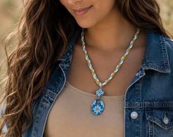 May include: A necklace featuring a braided cord in white, turquoise, and mustard yellow. The pendant consists of a small circular charm above a larger oval charm, both decorated with a blue and white floral mandala pattern on a silver base.