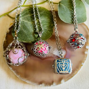 Puede incluir: Cuatro collares de plata con diferentes colgantes. Los colgantes son una jaula rosa y plateada con una bola rosa en el interior, una jaula rosa y plateada con un diseño de flor rosa, una jaula azul y plateada con un diseño de corazón y una jaula roja y plateada con joyas rojas y azules.