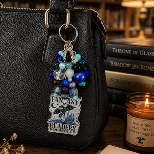 Puede incluir: Un bolso de mano de cuero negro con un llavero de cuentas. El llavero presenta un grupo de cuentas azules, transparentes y negras, y una etiqueta transparente con las palabras "Fantasy Readers Book Club" y una ilustración de dragón.