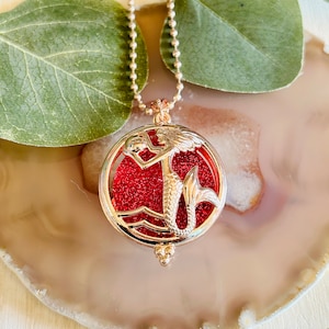 Könnte beinhalten: Eine goldene Kettenkette mit einem roten, mit Glitzer gefüllten Anhänger mit einem silbernen Meerjungfrauen-Motiv.