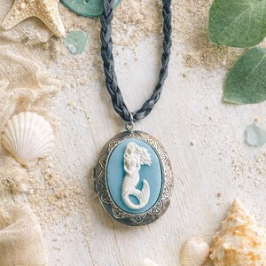 May include: A mermaid cameo pendant necklace with a braided gray cord. The oval pendant features a white mermaid on a light blue background, set in a silver-toned frame. Seashells and a starfish surround the necklace, evoking a beach theme.