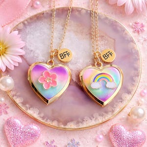 May include: Two gold necklaces with heart-shaped pendants. One pendant features a flower, the other a rainbow. Both pendants have a gold border and the letters "BFF" on a small charm. The background includes pink hearts and flowers.