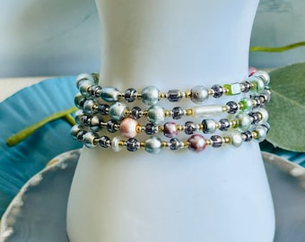 Puede incluir: Un brazalete de múltiples hilos con una mezcla de cuentas iridiscentes, plateadas y doradas. El brazalete está en capas con diferentes tamaños y colores de cuentas, creando un diseño único y llamativo.