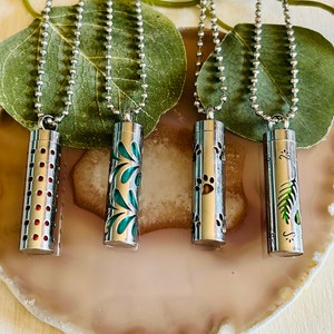 May include: Four silver necklaces with different designs on the pendant. The pendants are cylindrical and have a variety of patterns, including polka dots, leaves, paw prints, and a fern. The necklaces are on a light brown surface.