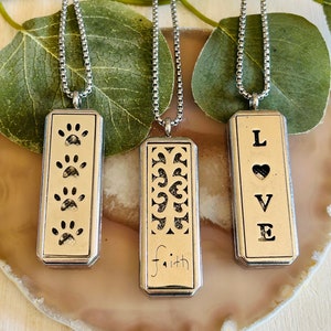 以下が含まれることがあります： 長方形のペンダントがついた銀のネックレスが3本。ペンダントには、足跡、装飾的なフィリグリー、そして「LOVE」という文字が刻まれています。フィリグリーペンダントには、「faith」という文字が下に刻まれています。