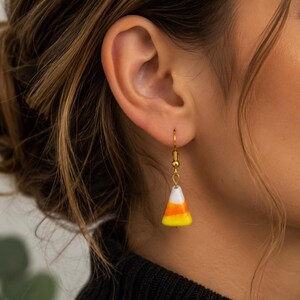 May include: A drop earring featuring a candy corn charm shaped like a triangular cone with horizontal bands of white, orange, and yellow. The charm hangs from a gold-toned metal hook that passes through the earlobe for secure placement.