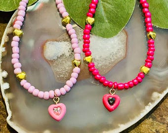 Puede incluir: Dos collares de cuentas con dijes en forma de corazón. Un collar es rosa con un dije de corazón rosa con un centro de cristal transparente. El otro collar es rojo con un dije de corazón rojo con un centro de cristal negro.