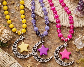May include: Three beaded necklaces with crescent moon and star pendants. The necklaces are yellow, purple, and pink. The pendants are silver-toned with yellow, purple, and pink stars. The beads are round and the necklaces are on a textured surface.