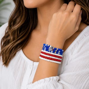 Puede incluir: Una pila de pulseras de cuentas con un patrón de colores azul, blanco y rojo. La sección azul incluye pequeñas cuentas metálicas y un dije de estrella plateado. Las cuentas son redondas y de tamaño uniforme, dispuestas en varias filas.