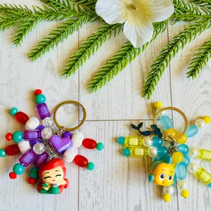 May include: Two beaded keychains featuring cartoon characters. One keychain has a purple heart charm and a red-haired mermaid figurine. The other keychain has a yellow and blue fish figurine and a blue whale charm. Both have gold key rings.