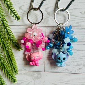Könnte beinhalten: Zwei Schlüsselanhänger mit Zeichentrickfiguren. Einer ist rosa mit einer Blume und rosa Perlen, der andere ist blau mit blauen Blumen und Perlen. Beide haben silberne Schlüsselringe und hängen an Haken.