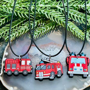 May include: Three red fire truck pendants on black cords. Each pendant features a different fire truck design with black, white, and gray accents. The pendants are displayed on a gray and white stone surface with green foliage in the background.