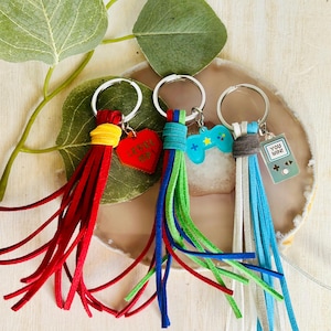 May include: Three colorful keychains with tassels. The first keychain has a red heart charm with the text "Level Up". The second keychain has a blue video game controller charm. The third keychain has a gray charm with the text "You Win" and a pixelated video game controller.