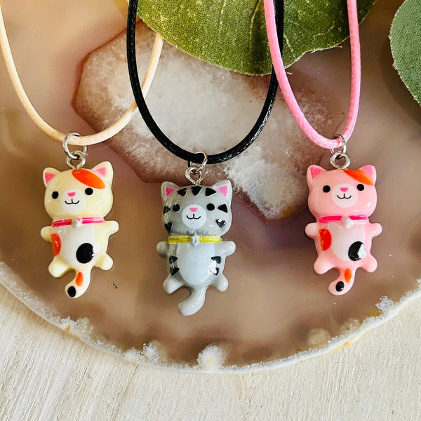 Pode incluir: Três colares com gatos de diferentes cores. Os gatos usam colares e têm olhos pretos. Os colares estão sobre uma superfície marrom clara.