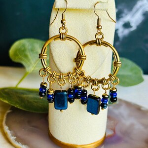 Può includere: Orecchini a cerchio dorati con perline blu e nere pendenti. I cerchi sono in filo metallico e hanno un design intrecciato. Le perline sono quadrate e rotonde.
