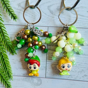 Könnte beinhalten: Zwei Schlüsselanhänger mit silbernen Ringen und Karabinerverschlüssen. Einer zeigt eine Zeichentrickfigur mit roten Haaren und einem grünen Hut, umgeben von grünen und braunen Perlen. Der andere hat eine blonde Zeichentrickfigur mit grünem Kleid und weißen Perlen.