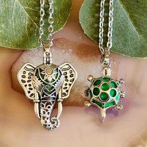 Könnte beinhalten: Zwei silberne Kettenhalsbänder mit Anhängern. Ein Anhänger ist ein Elefant mit grünen und silbernen Details. Der andere Anhänger ist eine Schildkröte mit einer grünen Schale.