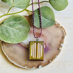 May include: A gold locket necklace with a burgundy chain. The locket has a striped design and is closed.