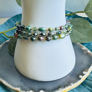 May include: A multi-strand bracelet with green, brown, and gold beads. The bracelet is made of different shapes and sizes of beads, including round, oval, and square beads.