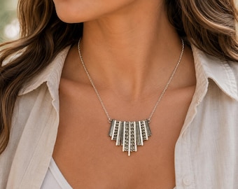 May include: A silver-toned necklace featuring a pendant composed of multiple vertical, textured metal bars arranged in a fan-like shape. The pendant is suspended from a delicate, thin silver-colored chain that rests around the neck area.