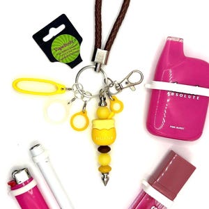 May include: A keychain with a brown braided strap, a yellow honey pot charm labeled HUNNY, and several silicone rings. Included are a pink disposable vape, a pink lighter, a white cylindrical tool, and a tube of pink lip gloss with a white silicone ring.