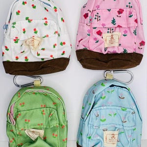 May include: Four miniature backpack keychains in white, pink, green, and light blue, each featuring a floral pattern, a brown base, a front zippered pocket, a top zipper, and a silver metal carabiner clip. Each bag has a small rectangular label.