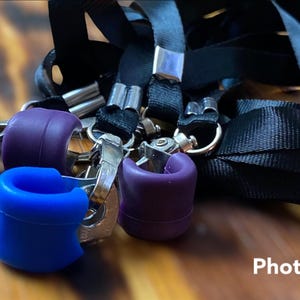 May include: Close-up view of several lanyard attachments with various colored silicone end caps.  The caps are in shades of purple and blue, and black.  Silver metal hardware is visible on each attachment.  The lanyards themselves are black. The image includes the watermark 'PhotoRoom®'.