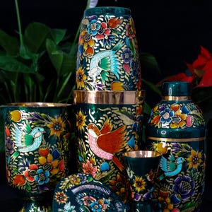 May include: A collection of hand-painted, decorative ceramic items. The pieces include a cocktail shaker, glasses, and a hand mirror. Each item is dark green with colorful floral and bird designs, accented with gold trim.