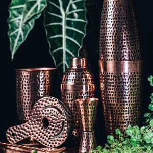 Pode incluir: Uma coleção de ferramentas de bar de cobre, incluindo uma coqueteleira, um jigger, um coador e dois copos de tamanhos diferentes. As ferramentas têm uma textura martelada com pequenos orifícios espaçados uniformemente. As ferramentas de cobre são dispostas em frente à folhagem verde.