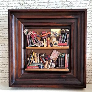 Może przedstawiać: Miniaturowa biblioteczka z ciemnobrązową drewnianą ramą, wypełniona małymi książkami. Książki są ułożone na dwóch półkach i mają różne tytuły, w tym "LOVE" i "AGGEZZA".