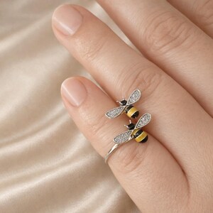 May include: A silver-toned open ring featuring two bee motifs. Each bee has wings encrusted with small clear stones, a black head, and a body with alternating yellow and black stripes. The ring band is thin and metallic, wrapping around the finger.