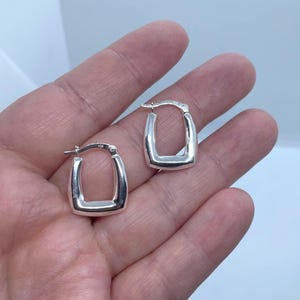 May include: A pair of silver-coloured square hoop earrings. The earrings have a smooth, polished surface and a rectangular shape with rounded corners. The earrings are displayed against a light background.