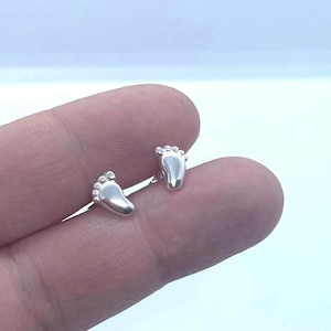 Puede incluir: Un par de pendientes plateados con forma de pies pequeños. Los pendientes están muy pulidos y reflejan la luz, con detalles que muestran los dedos de los pies. El fondo es azul claro.