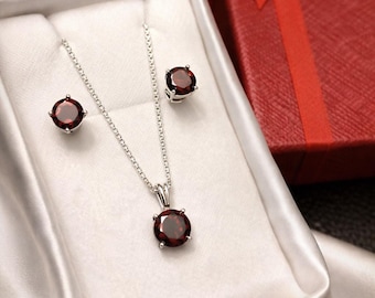 May include: A jewellery set consisting of a silver-toned chain necklace with a round, dark red gemstone pendant and a pair of matching round, dark red gemstone stud earrings, all presented in a white satin-lined box with a red gift box nearby.