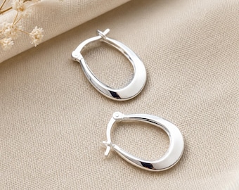 May include: A pair of polished silver hoop earrings with a smooth, elongated oval shape. The earrings feature a hinged post mechanism at the top, designed for secure closure. The metal surface is reflective and uniform, showcasing a clean, metallic finish.