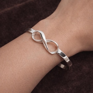 May include: A polished silver-toned cuff bracelet featuring a central infinity symbol design. The metal band is smooth and reflective, curving around the wrist to form a continuous loop shape that sits prominently at the centre of the bracelet.