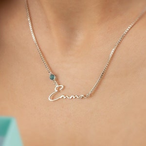 May include: A silver necklace spelling "Emma" in cursive. The necklace has a small, blue gemstone and is worn around the neck. A light blue gift box is being held.