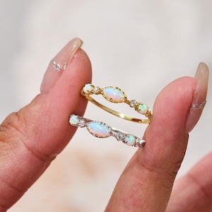 May include: Two delicate rings with oval-shaped opal stones set in gold and silver. The rings are stacked on a finger.