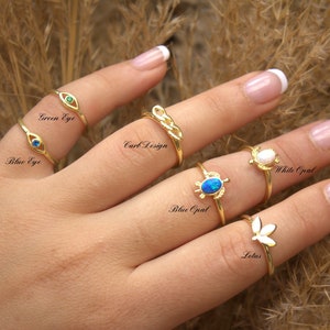 May include: A hand displays several gold rings with various designs. Rings include an eye with a green stone, an eye with a blue stone, a knot design, a turtle with a blue opal, a white opal flower, and a lotus flower. Text on the image labels each ring.