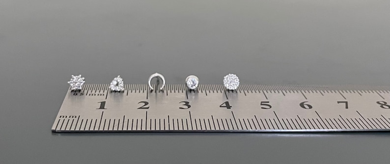 May include: Five different silver nose rings with clear rhinestones, measured on a ruler in inches.