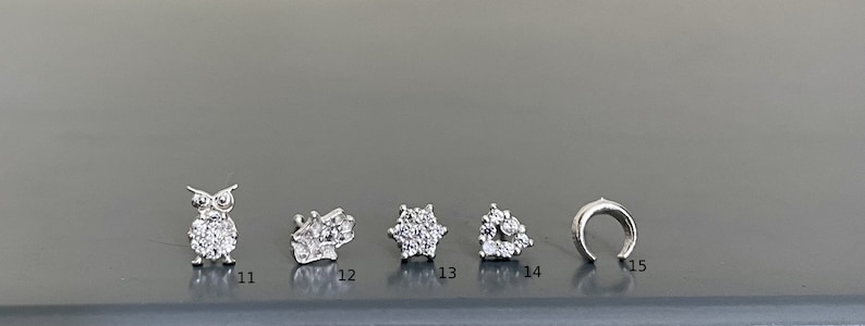May include: A set of six silver nose rings with different designs, including an owl, a flower, a star, a heart, and a hoop. The rings are all set with clear cubic zirconia stones.
