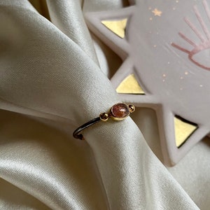 May include: A gold ring with a pink and brown oval gemstone set in the center. The ring is on a white satin fabric background.